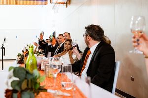 Wedding party guests raising glasses in a toast at the head table decorated with greenery.