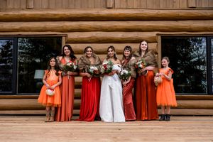 Wedding party in orange and red dresses poses together on wooden deck of rustic log cabin venue.