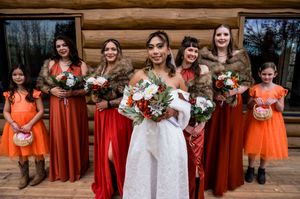 Wedding party in orange and red dresses stands together in front of log cabin.