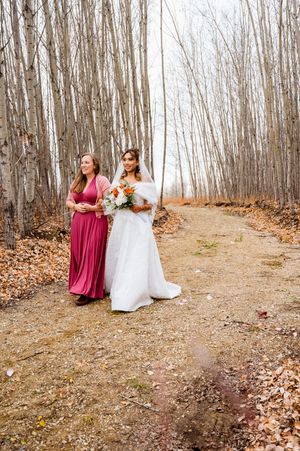 Wedding party walking through a forest path lined with bare trees and fallen autumn leaves.