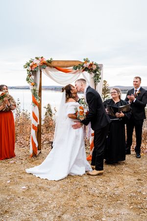 An outdoor wedding ceremony featuring orange and white decor with guests gathered on a cloudy autumn day.