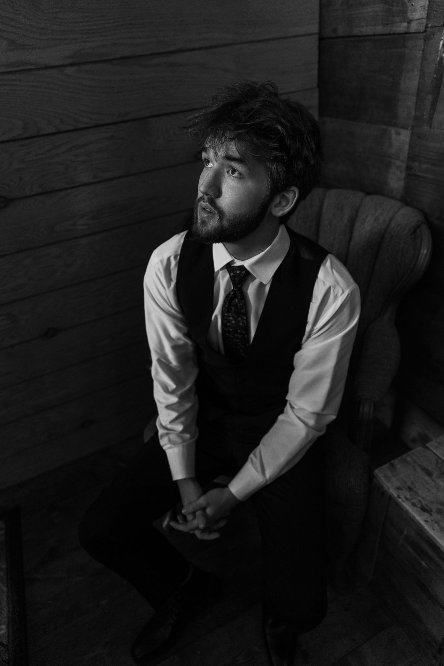 Black and white portrait of someone in formal attire with vest and tie sitting thoughtfully in dark wooden room.