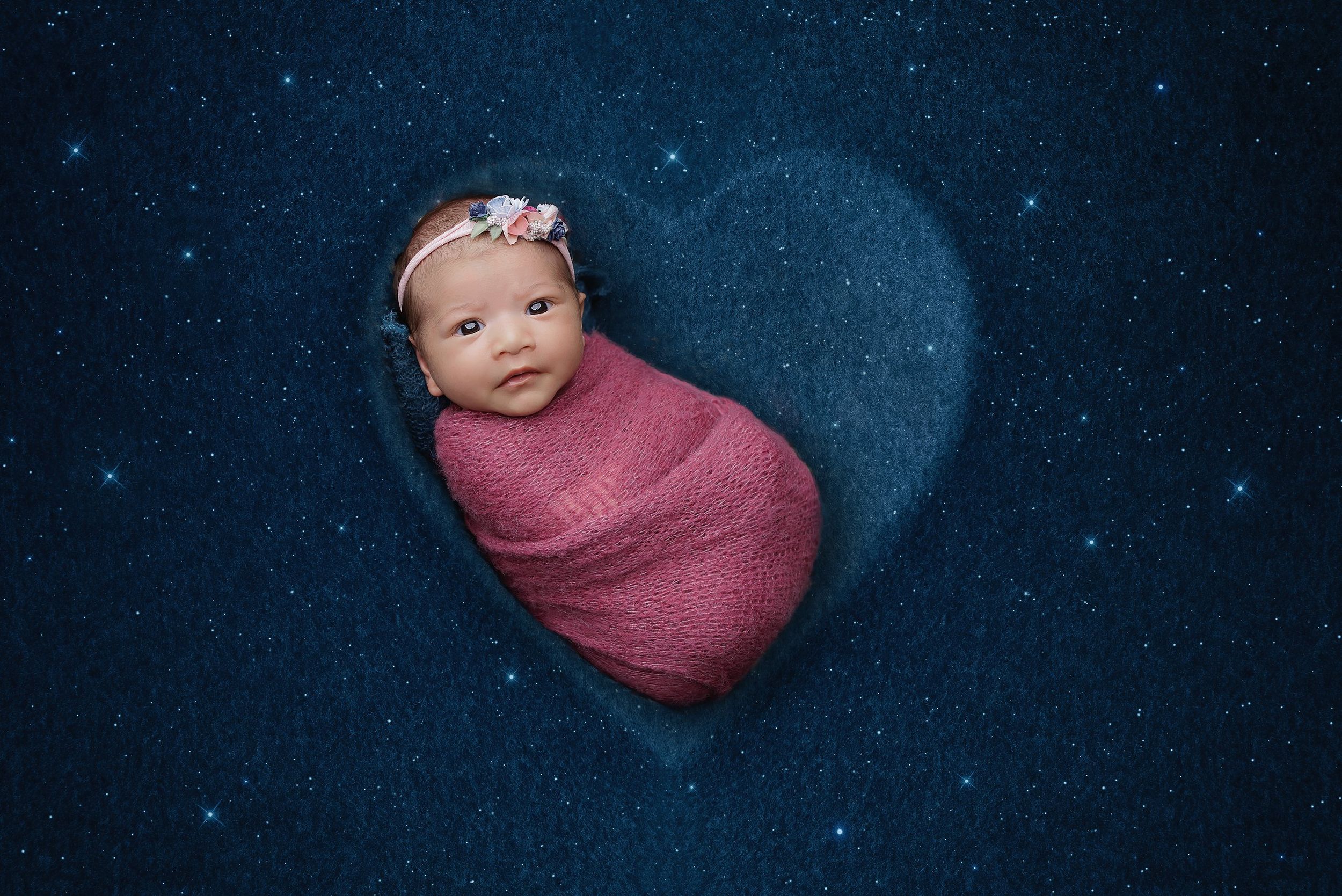 Newborn girl swaddled on navy blue starry night backdrop with heart shape