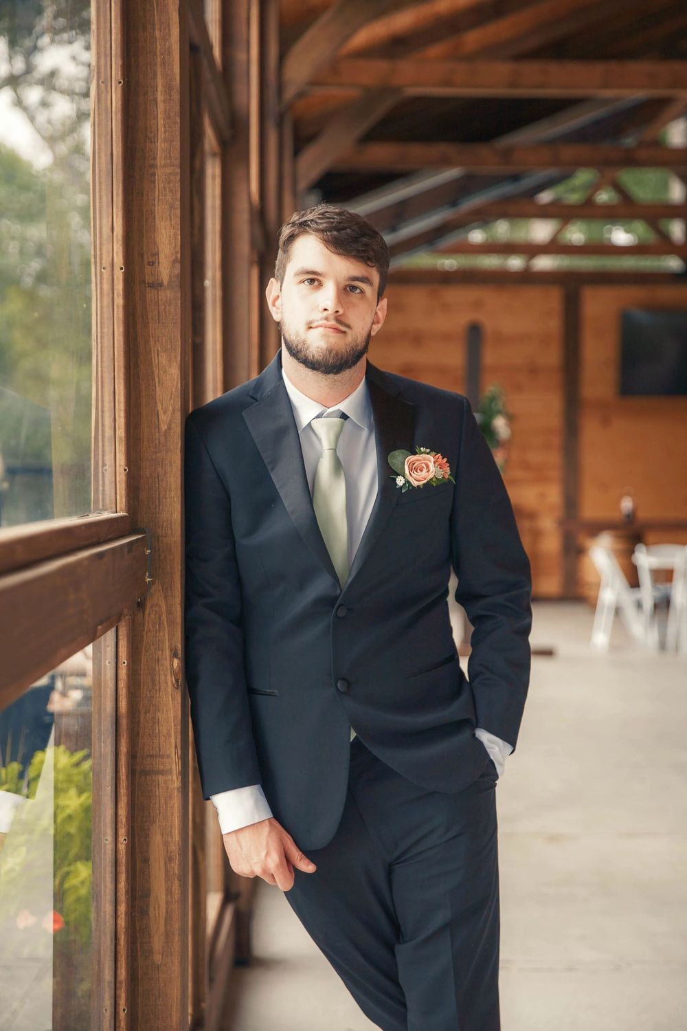 Handsome groom in navy suit with floral boutonniere leaning against wooden wall at wedding venue.