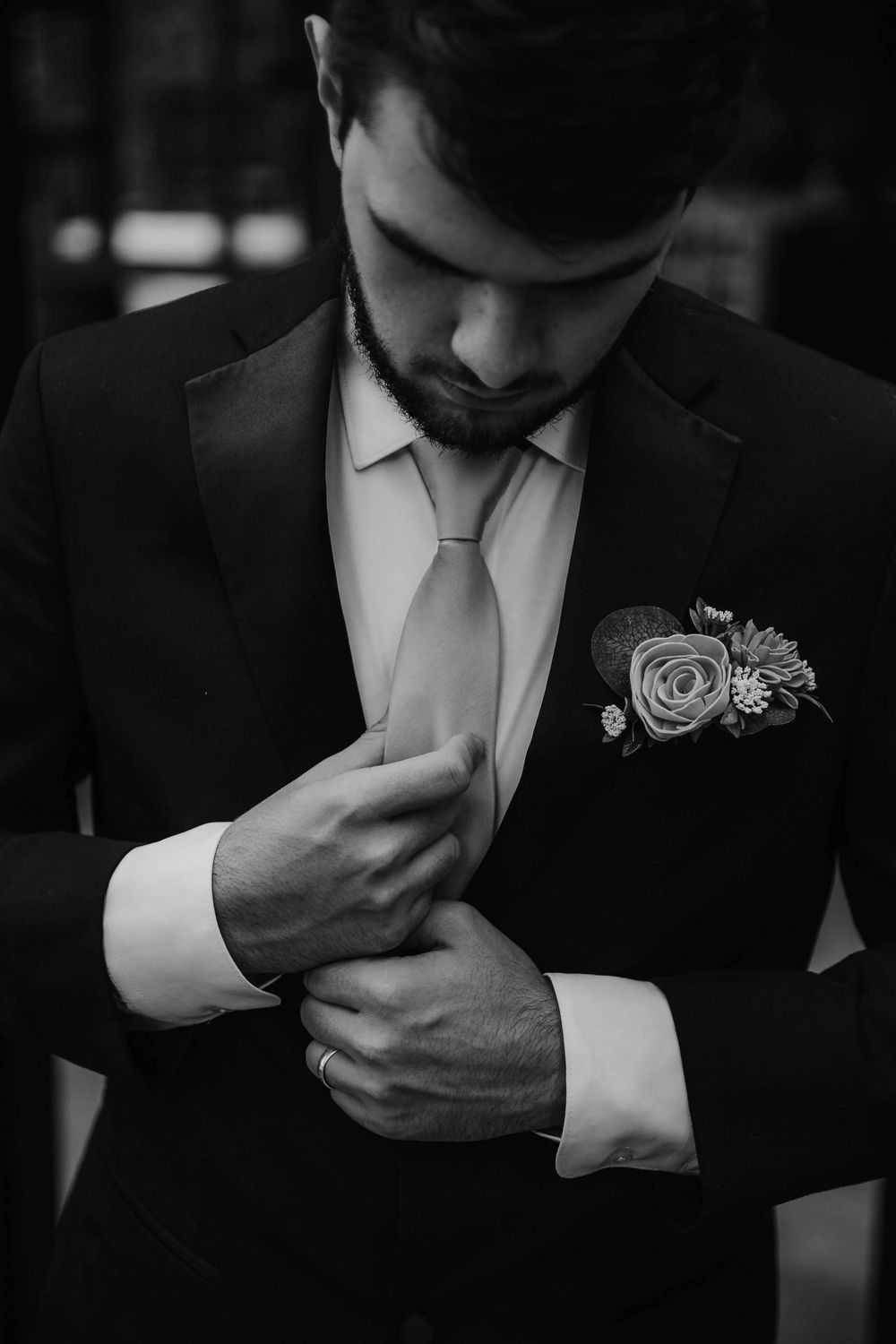 Black and white portrait of dapper groom in suit adjusting cufflinks with boutonniere on lapel.