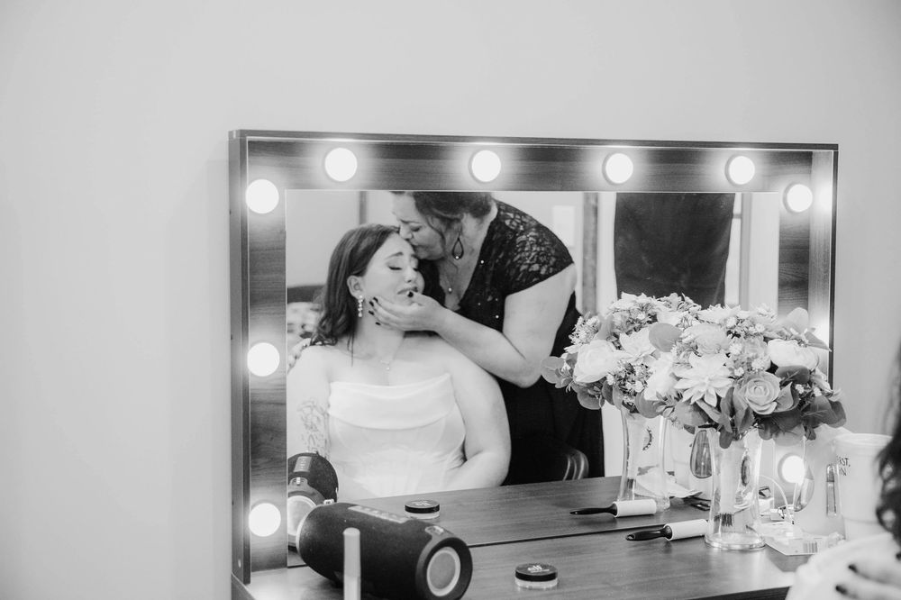 Black and white photo of bride getting makeup applied at vanity mirror with Hollywood lights.