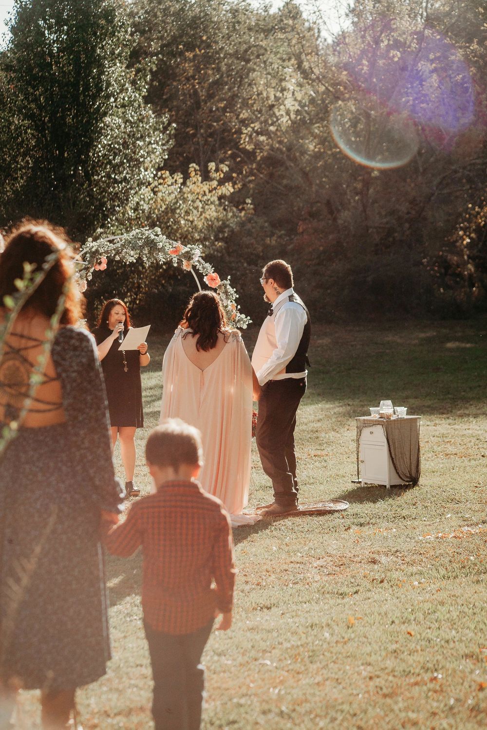 Outdoor wedding ceremony at golden hour with bride and groom standing beneath floral arch surrounded by guests.