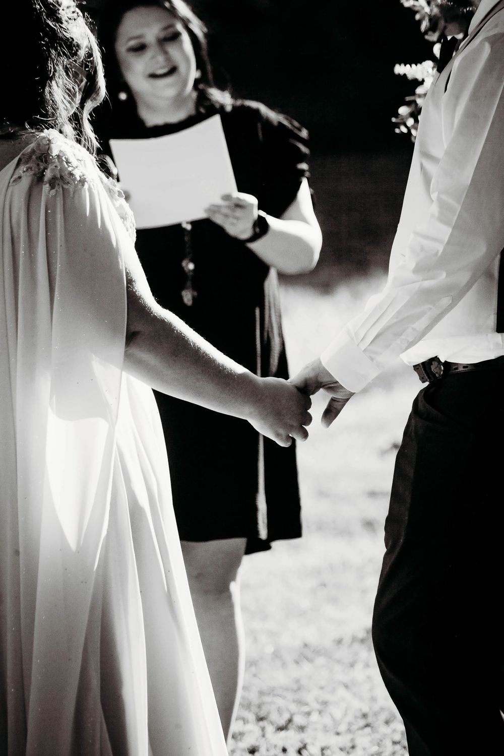 Black and white photo of bride and groom holding hands during outdoor wedding ceremony with officiant reading vows.