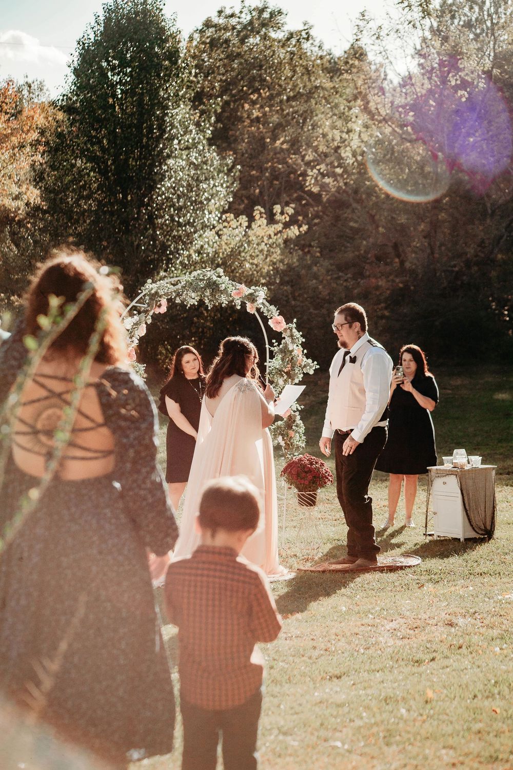 Bride and groom exchange vows under floral arch at sunny outdoor wedding ceremony with guests watching.