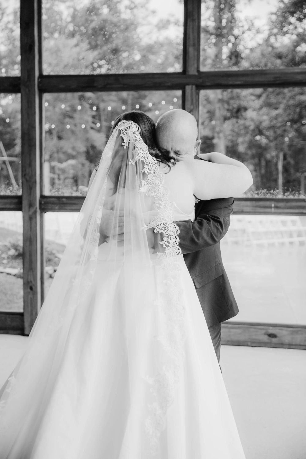 Black and white photo of bride and groom sharing emotional first look embrace by large window.
