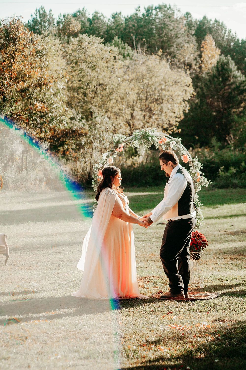 Bride and groom holding hands under floral arch in sunny outdoor wedding ceremony.