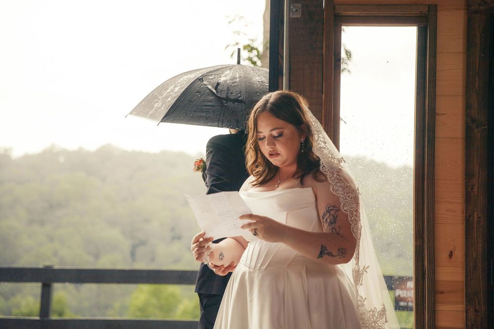 Bride in lace veil reads letter while groom holds umbrella during rainy wedding day first look.