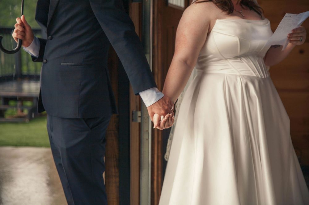 Bride and groom hold hands through doorway during rainy outdoor wedding day first look moment.