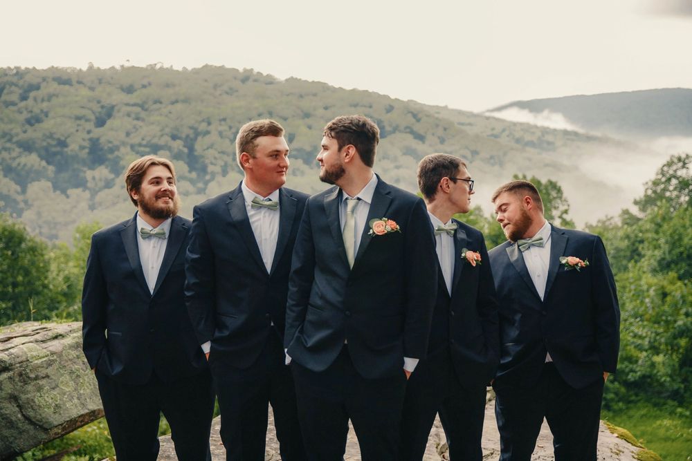 Five groomsmen in navy suits and bow ties laughing together outdoors with misty mountain views.