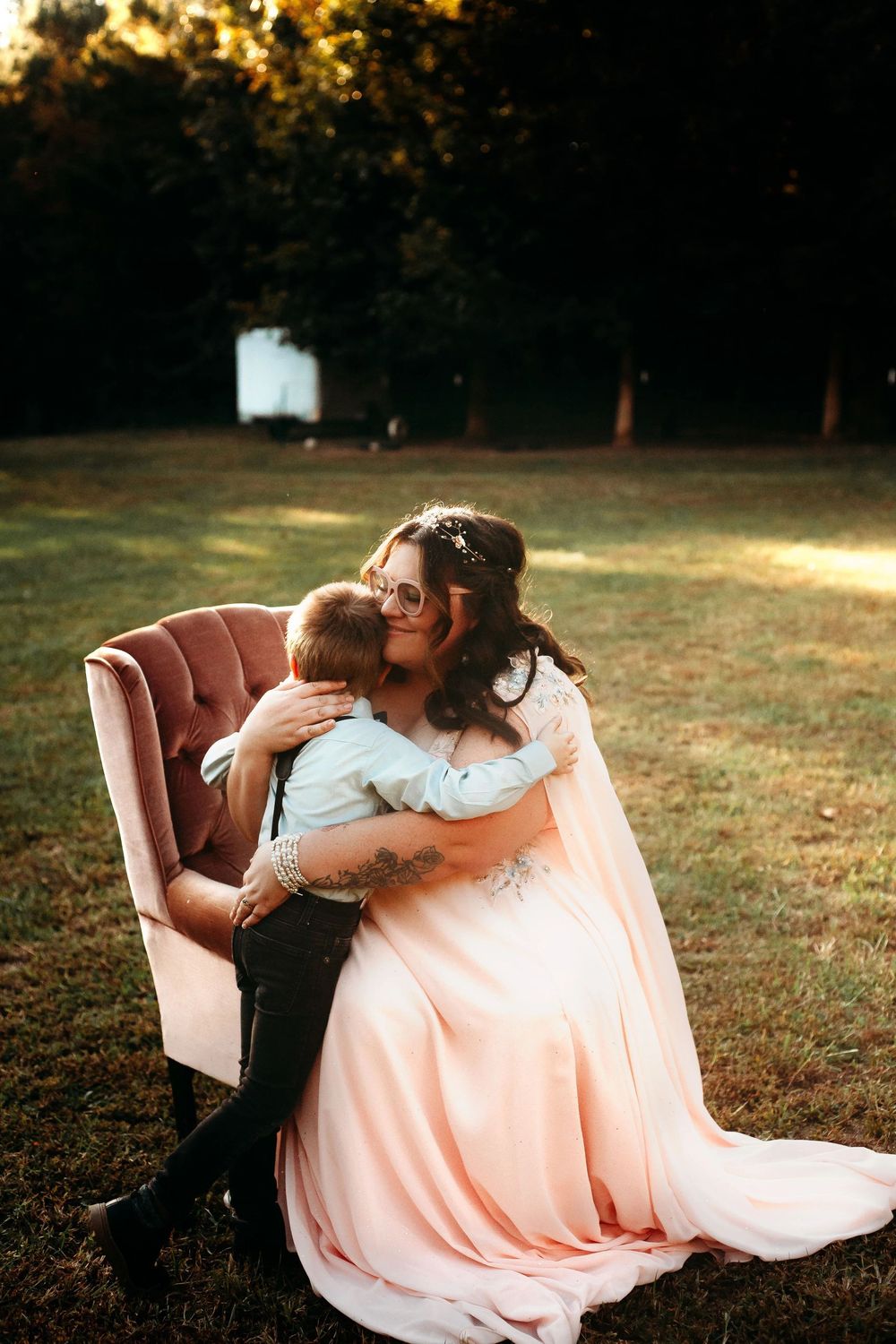 Bride in pink gown embracing young boy in velvet chair at outdoor wedding.
