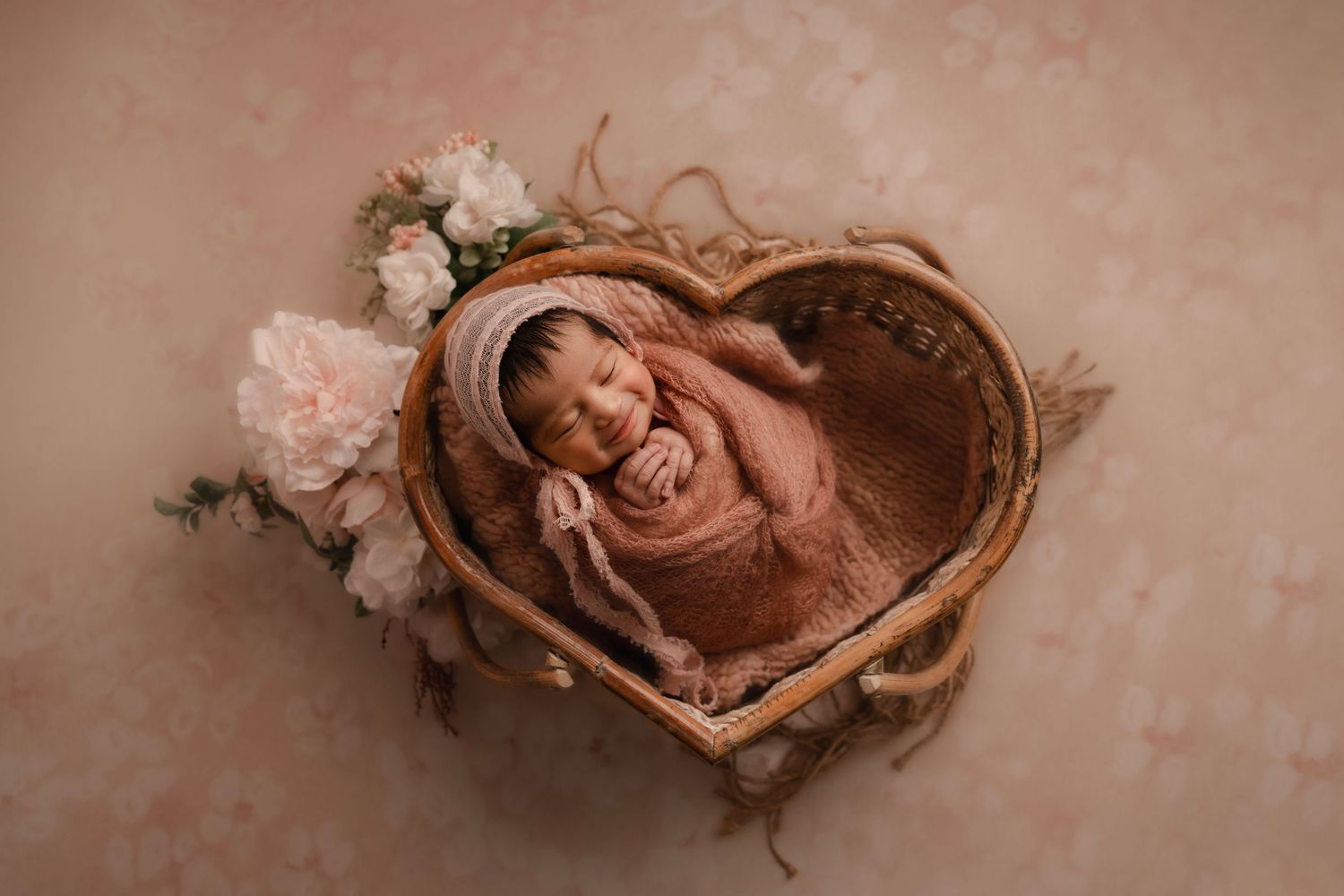 Newborn baby smiling, swaddled in pink wrap inside a heart-shaped wicker basket with flowers.
