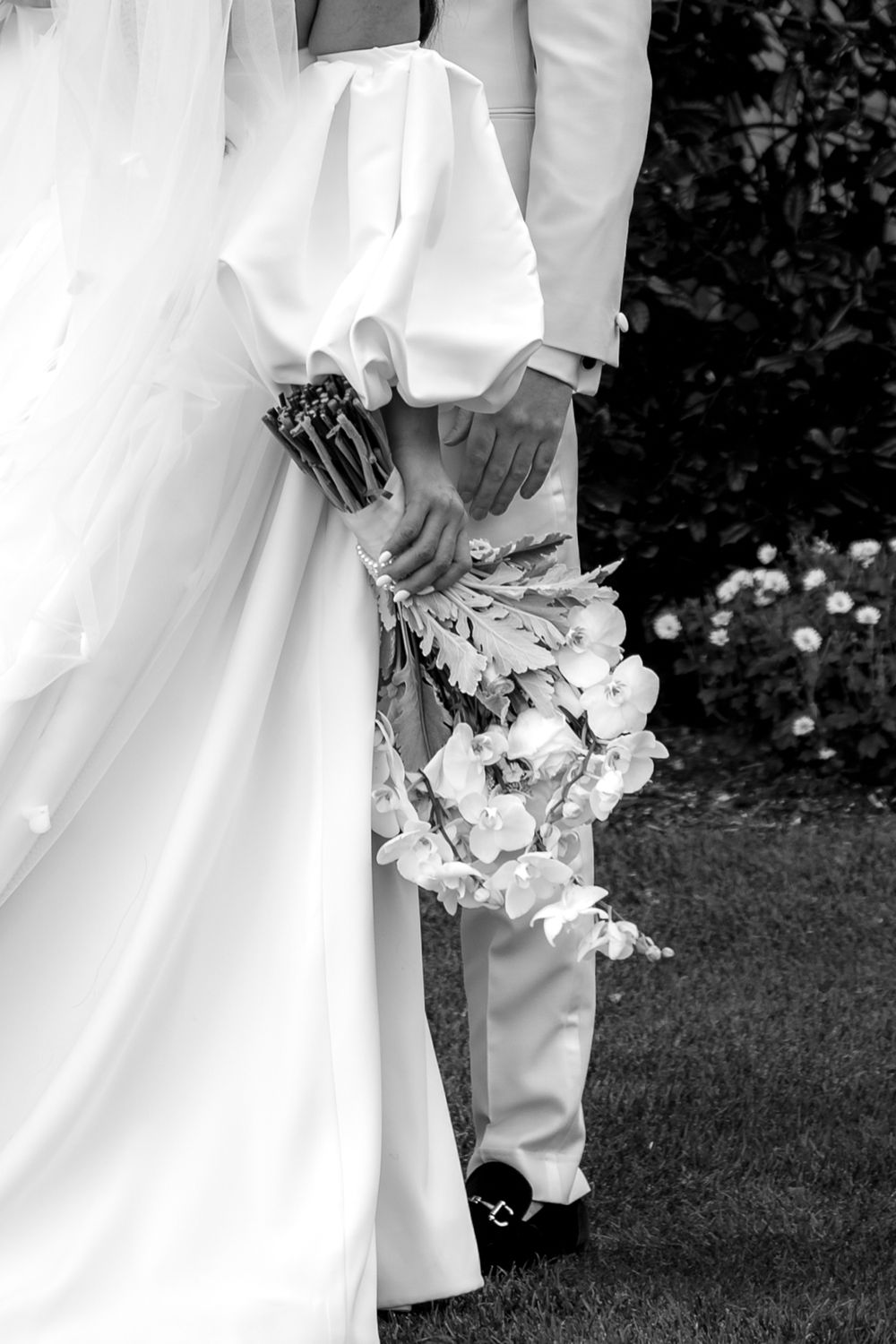 A couple in white wedding attire stands together holding a bouquet of white flowers on a grassy lawn.