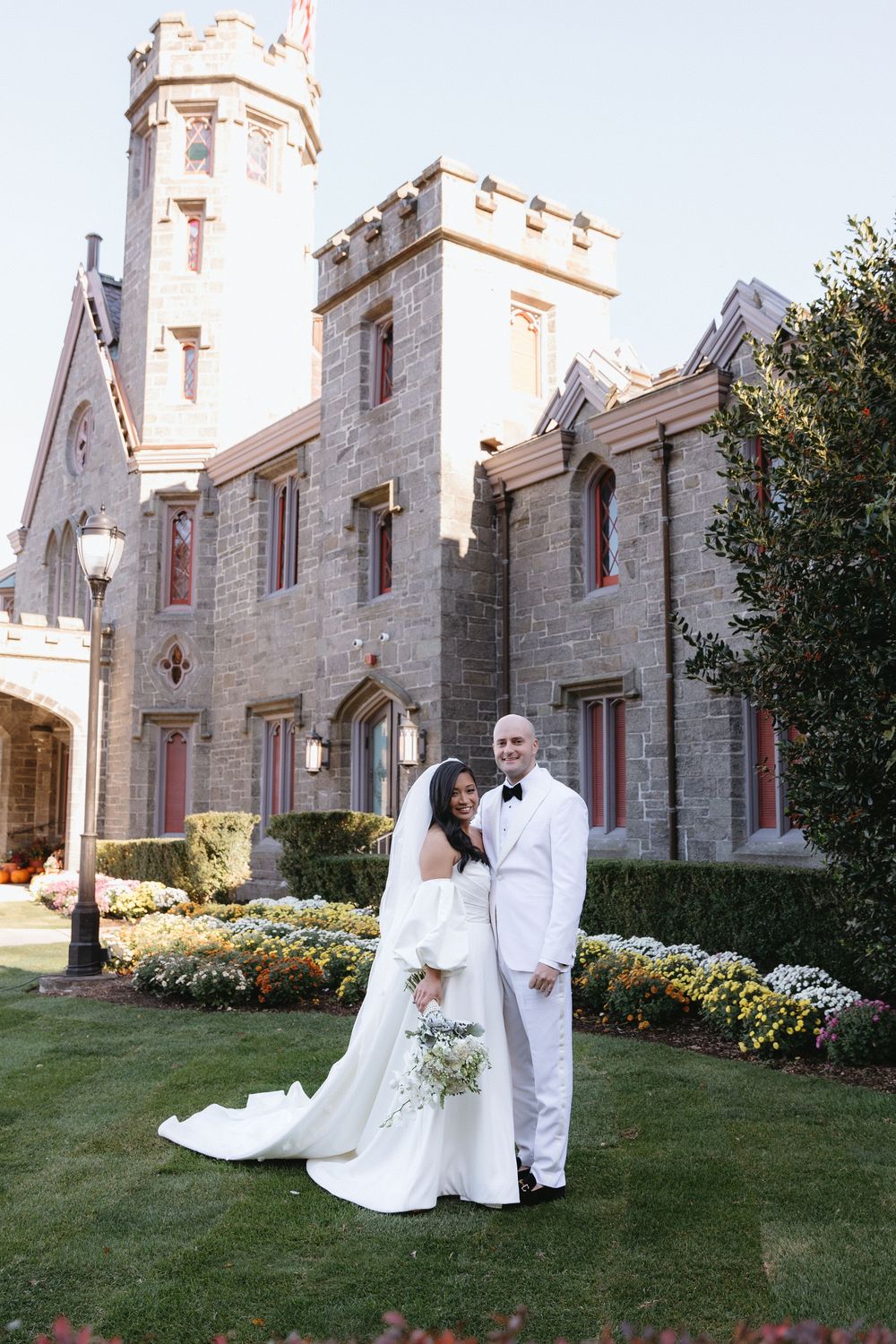 Elegant wedding couple poses in front of a stone gothic church with beautiful landscaping.