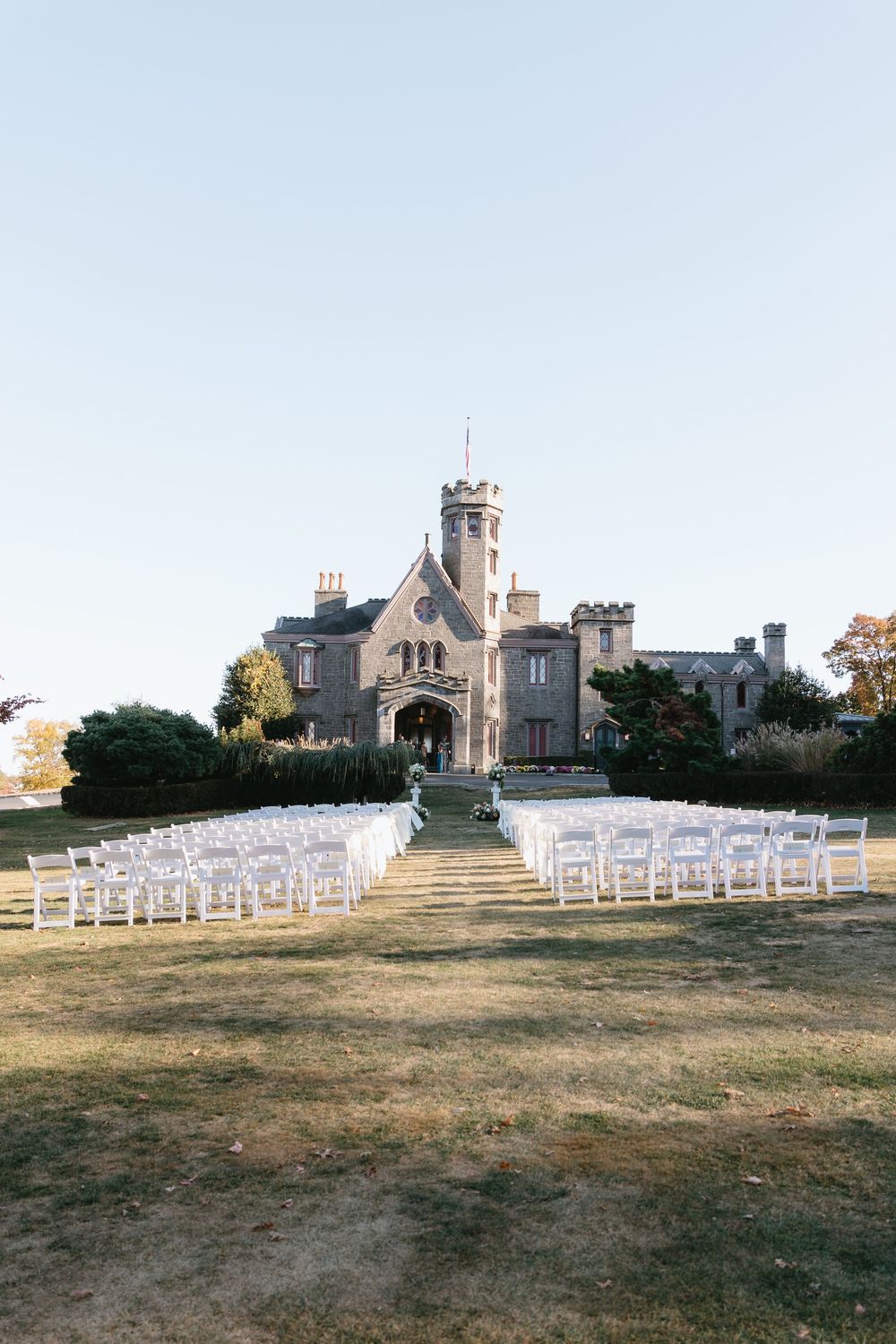 White chairs arranged for an outdoor wedding ceremony with a stone castle-like building in the background.