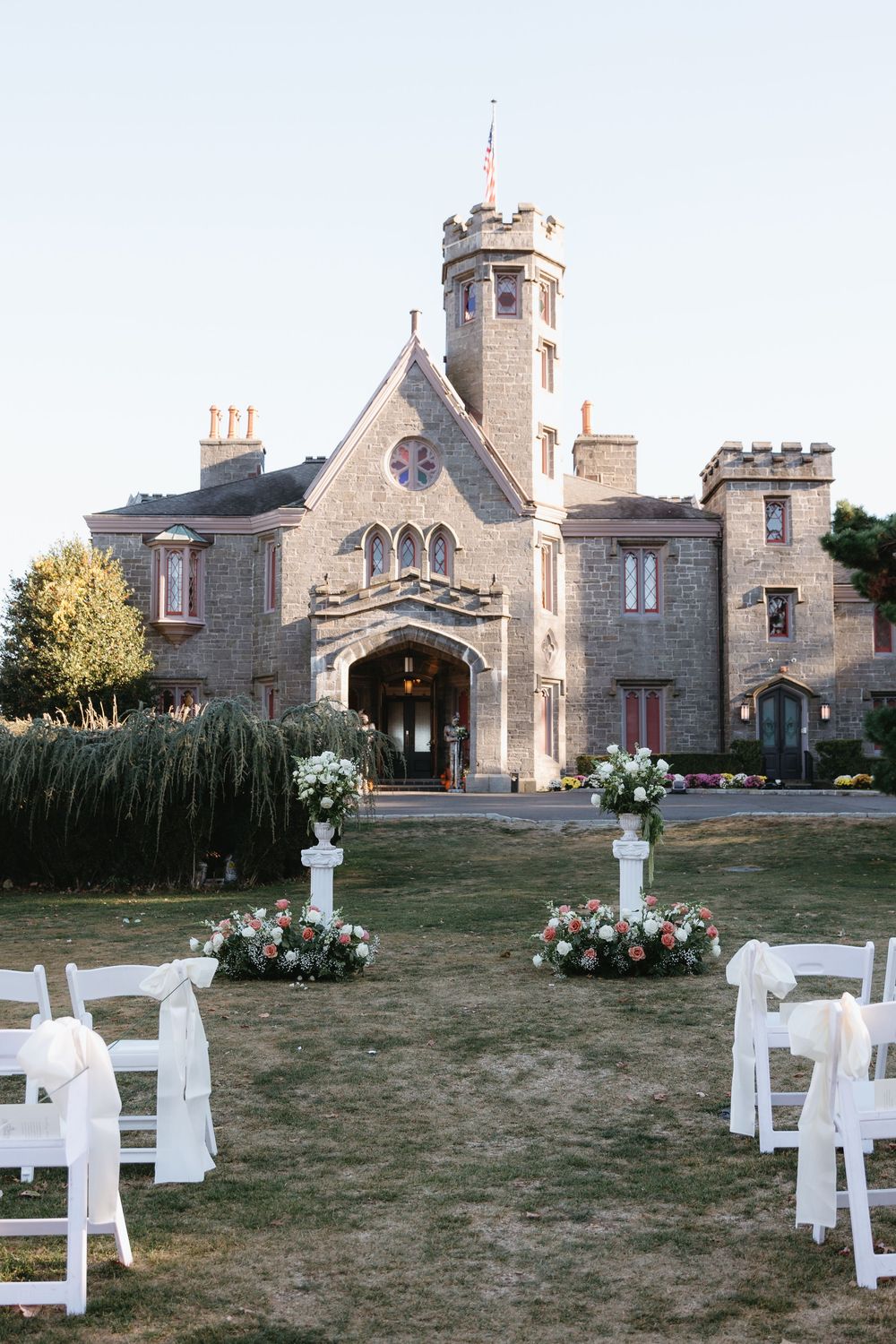 Elegant outdoor wedding ceremony setup with white chairs and floral arrangements in front of a stone castle building.