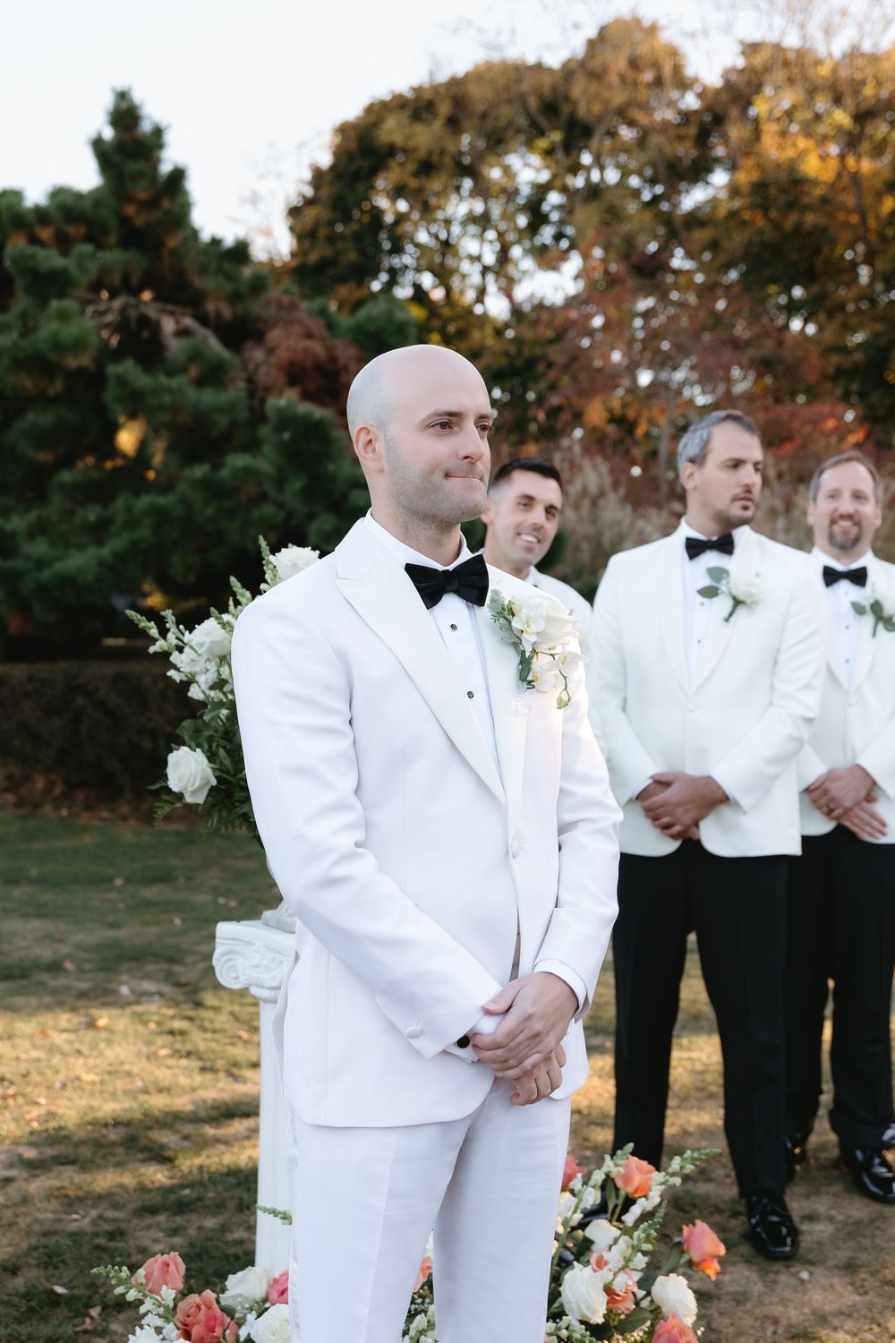 Wedding party members in crisp white tuxedos and black bowties stand together during an outdoor autumn ceremony.