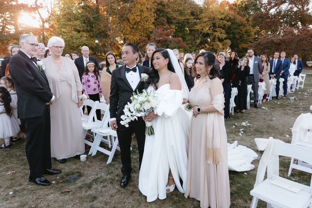 Wedding guests gather outdoors during an autumn ceremony with fall foliage and white attire.