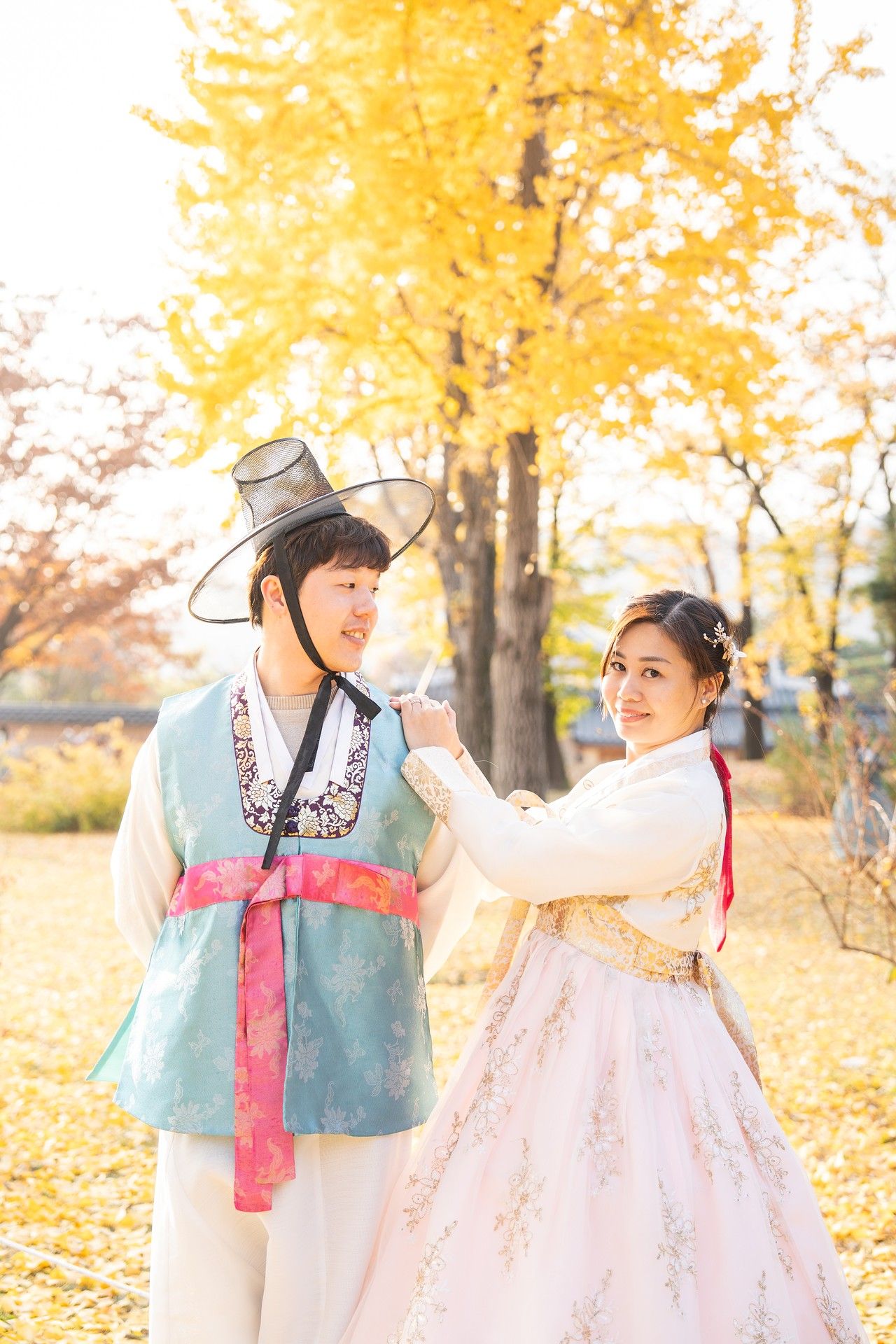 A couple in traditional Korean hanbok outfits pose together in a park with golden autumn leaves in the background.