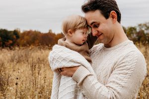 A family wearing cozy cream sweaters embraces tenderly in a golden autumn field at sunset.
