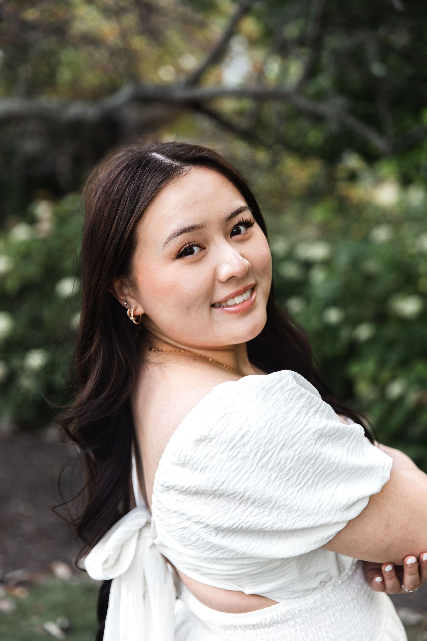 A person in a white blouse poses playfully against a green outdoor background.