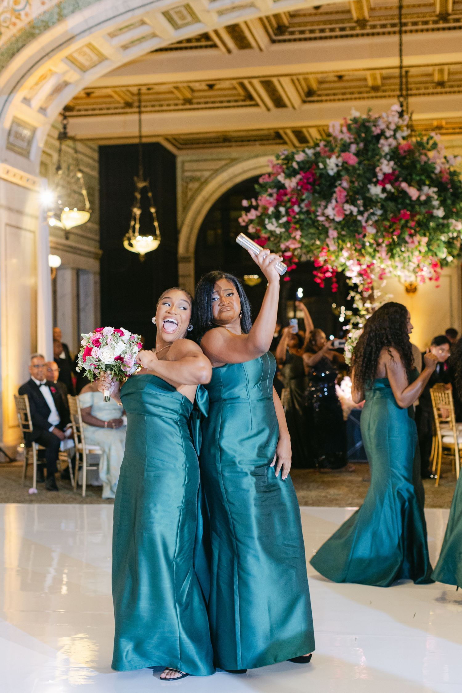 Bridesmaids in teal satin gowns celebrate at an elegant ballroom reception with ornate gold ceiling and floral arrangements.