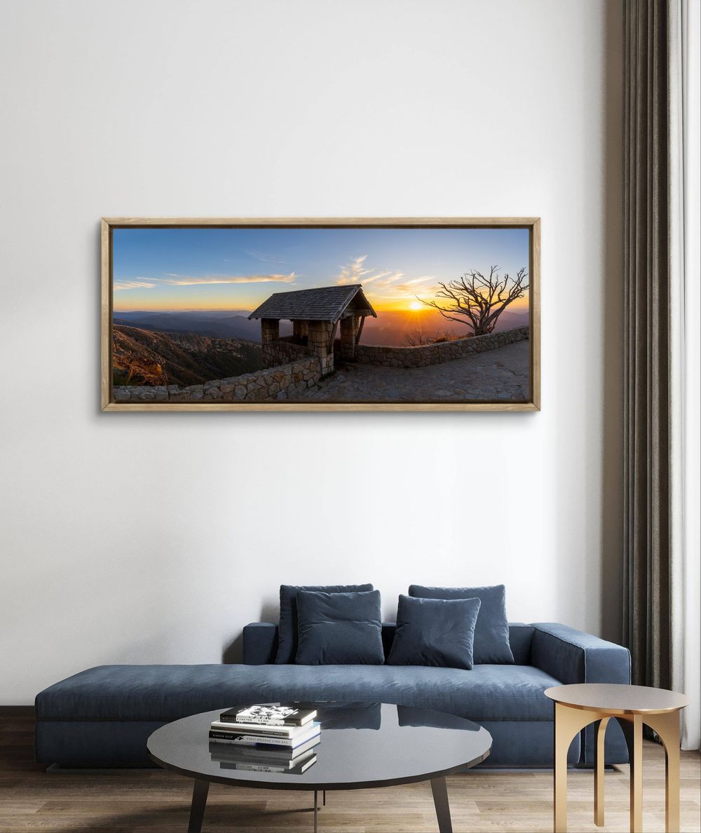 Large framed panoramic sunset print with rustic hut and bare tree displayed above a blue sofa.