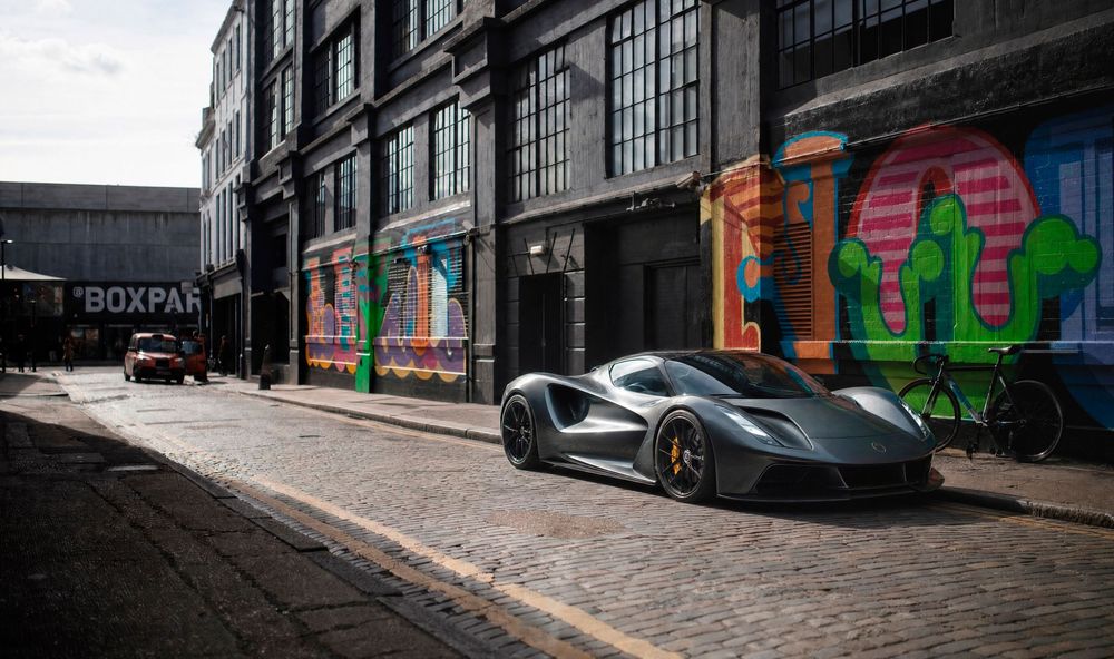 A sleek gray Lotus Evija electric hypercar parked on cobblestone street beside colorful murals in an urban setting.