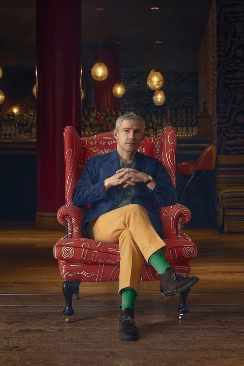 A person wearing yellow pants and green socks sits in a red velvet armchair with warm lighting in the background.