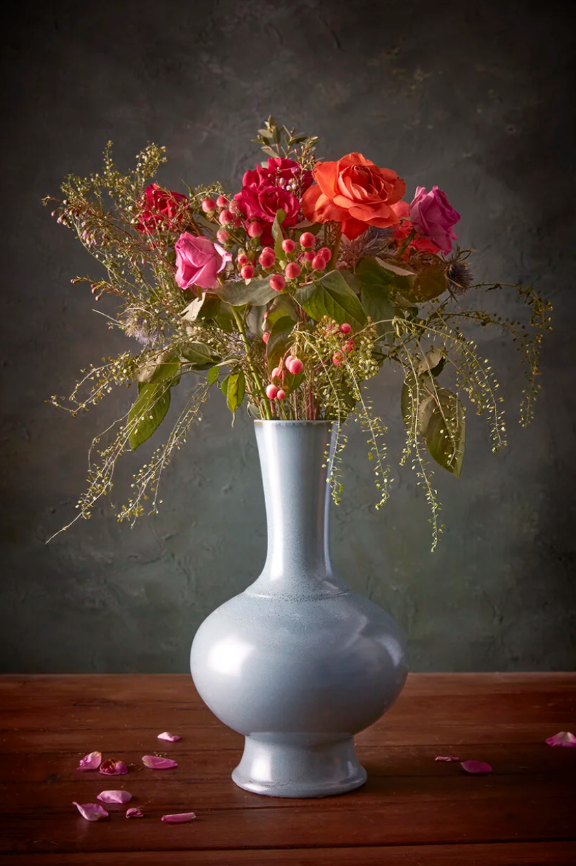 A dramatic still life of red roses and flowers arranged in a light gray ceramic vase against a dark background.