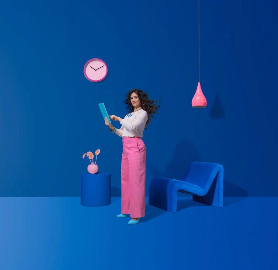 Person in pink dress stands against blue wall while geometric shapes create dynamic composition and shadows.