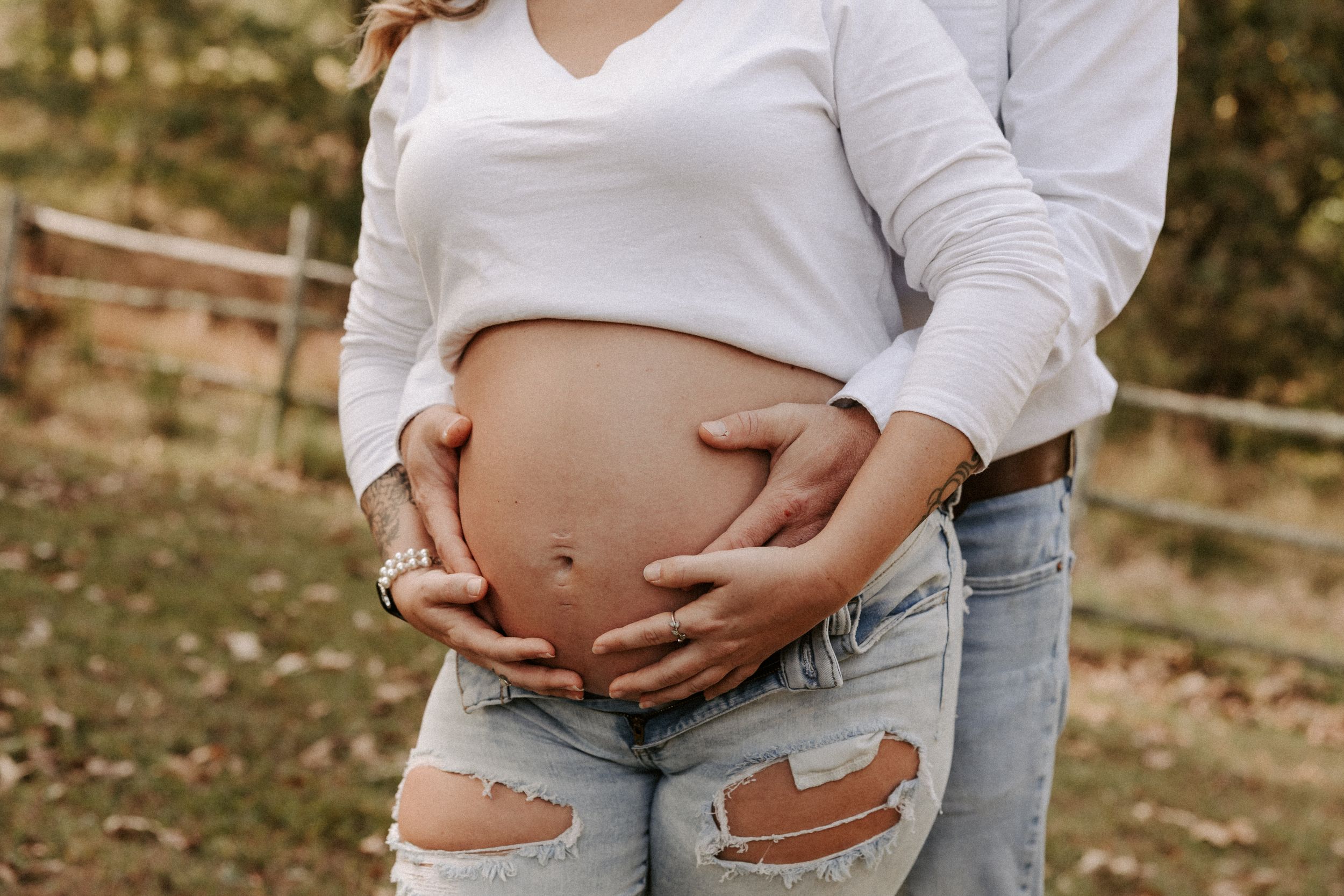 A casual maternity photo showing hands cradling a baby bump, wearing distressed jeans and a white crop top.