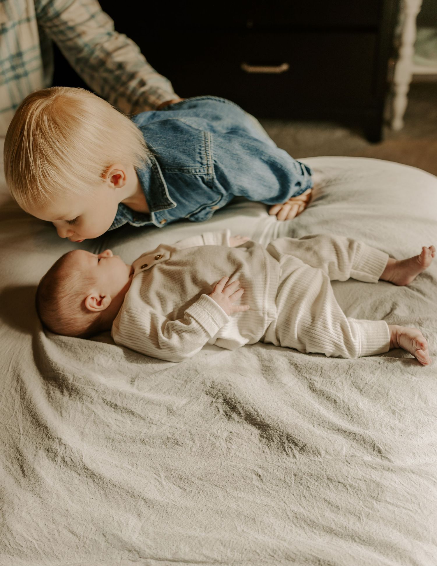 Side view of baby resting on white bedding with sibling nearby.
