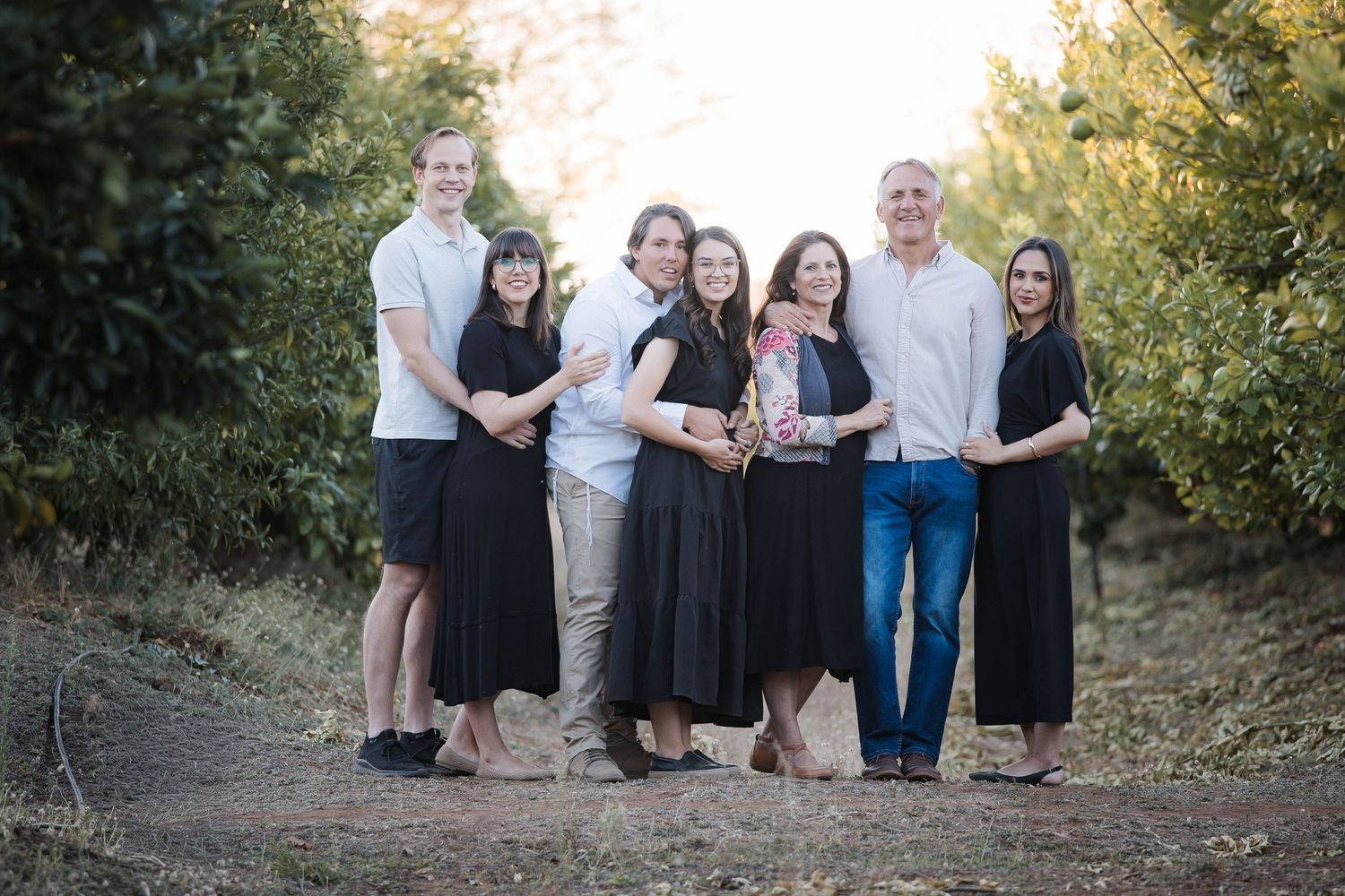 A group of people pose together outdoors during golden hour in a natural setting with greenery in the background.