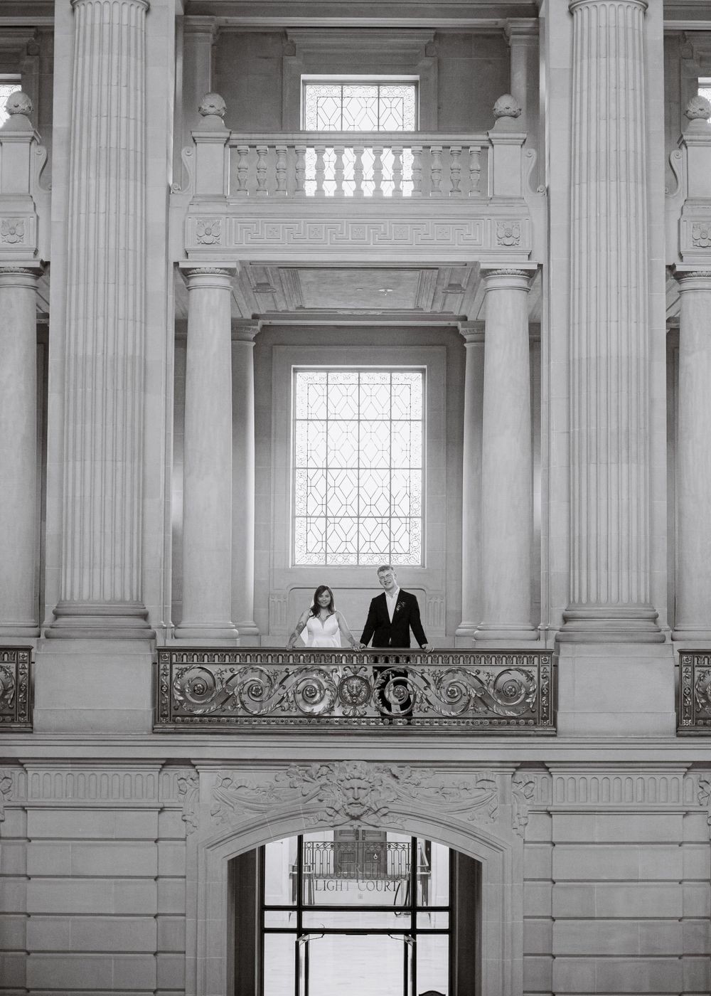 A couple stands on an ornate balcony inside a classical building with towering columns.