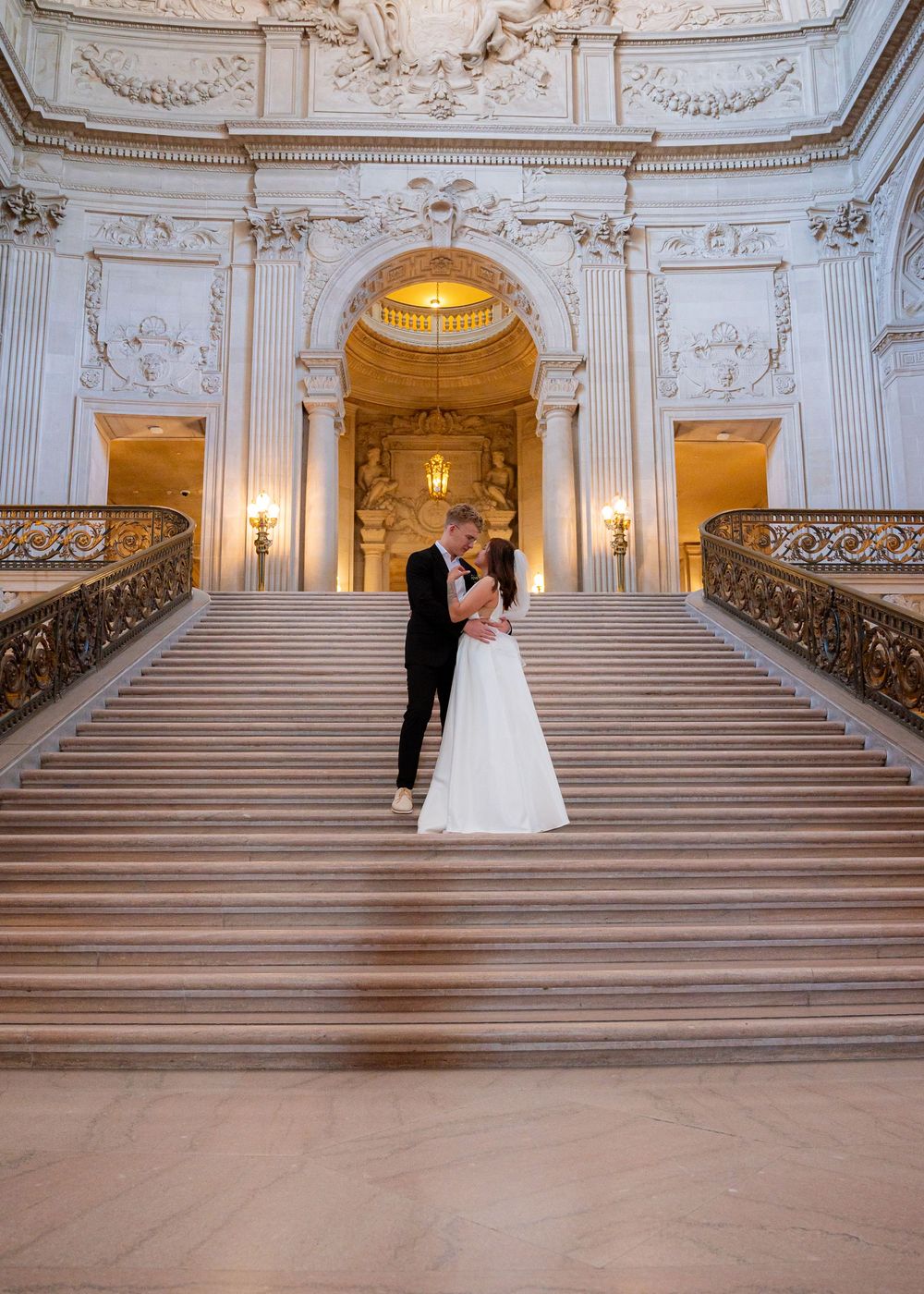 A grand marble staircase with ornate baroque details and glowing wall sconces in San Francisco City Hall's rotunda.