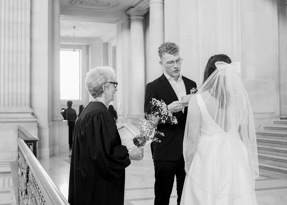 A black and white photograph of a wedding ceremony taking place in an elegant hallway with classical columns.