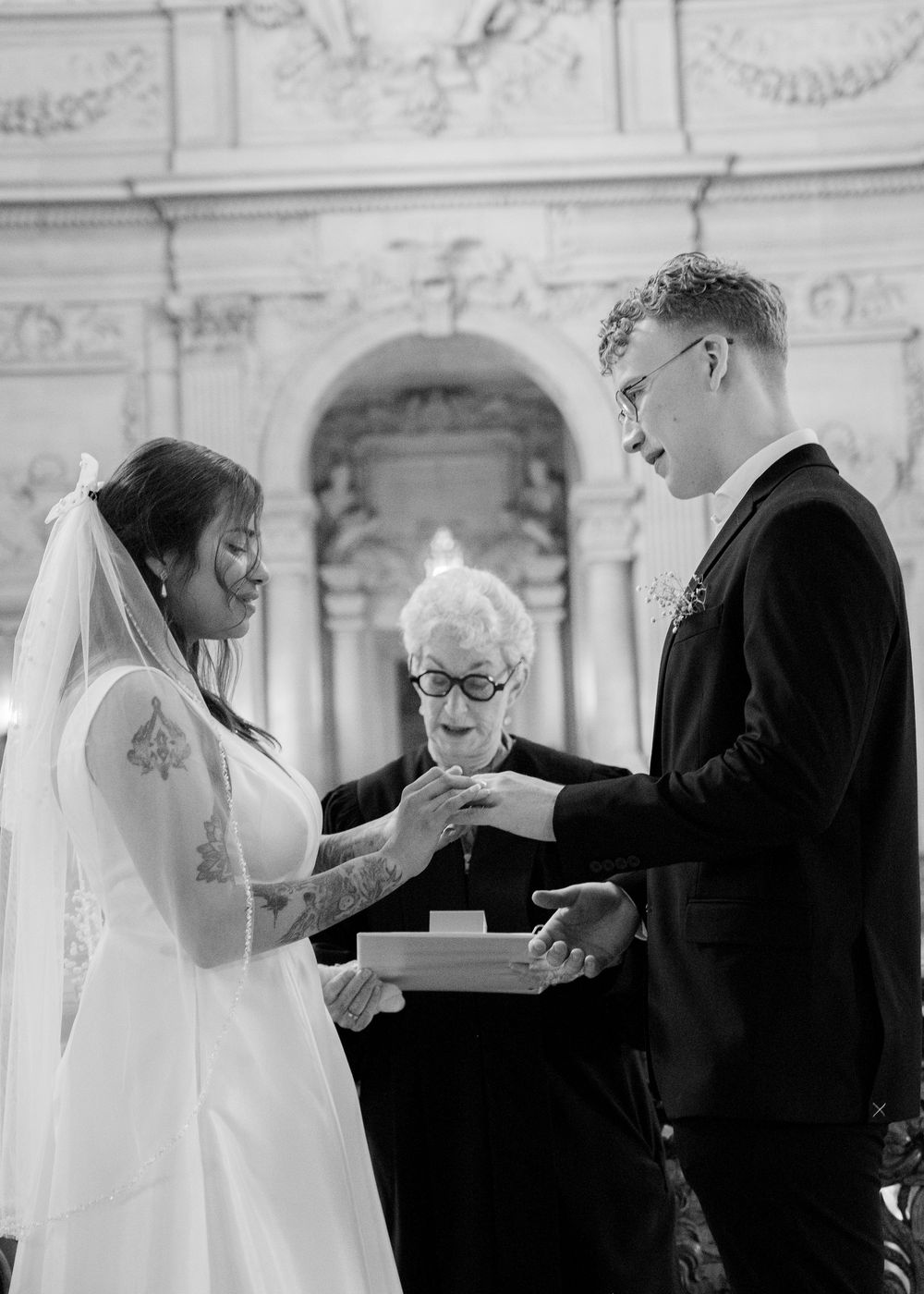 A couple exchanges rings during their wedding ceremony with an officiant in a grand architectural setting.
