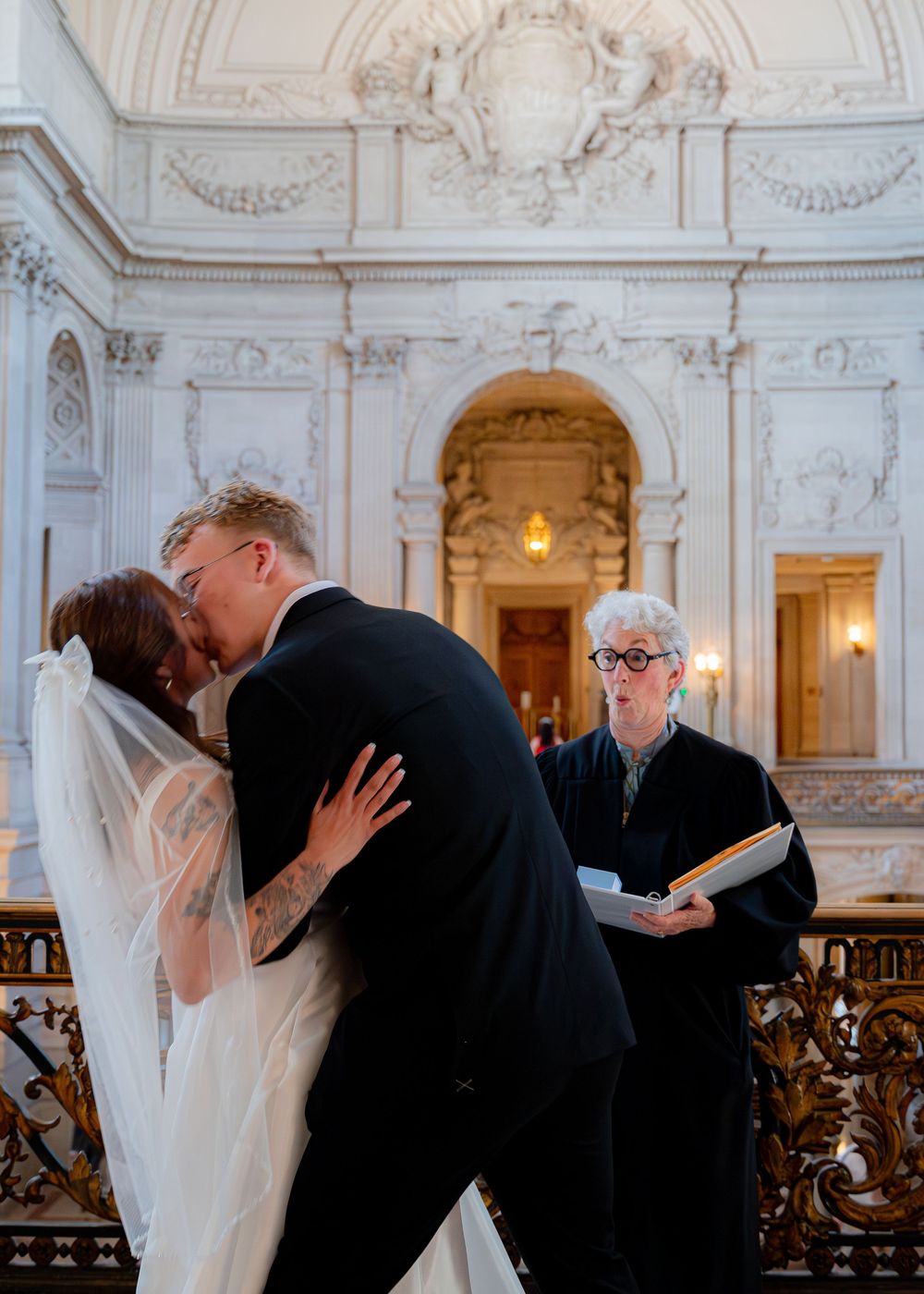 A wedding kiss moment captured in an elegant historic building with ornate white architectural details.
