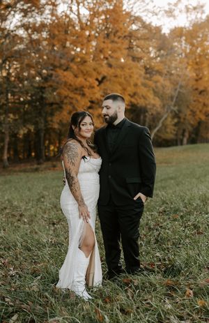 Wedding couple stands together in a scenic fall field with orange-leaved trees in background.