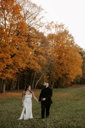 Wide landscape wedding shot with couple walking through field beneath orange autumn trees.