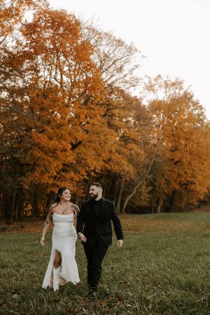 Wedding couple walks hand in hand through meadow with autumn foliage in background.