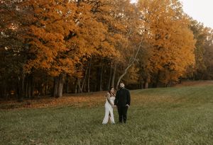 Distant view of newlyweds walking together in a grassy field with fall trees.