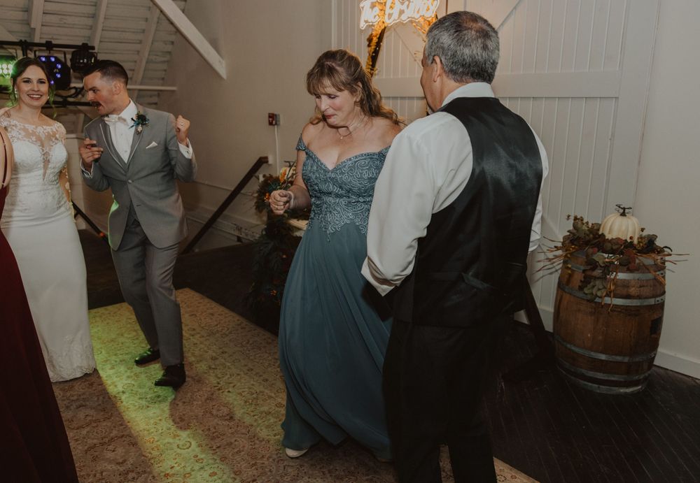 Wedding guests enjoying themselves on a dance floor in a rustic indoor venue with string lights.