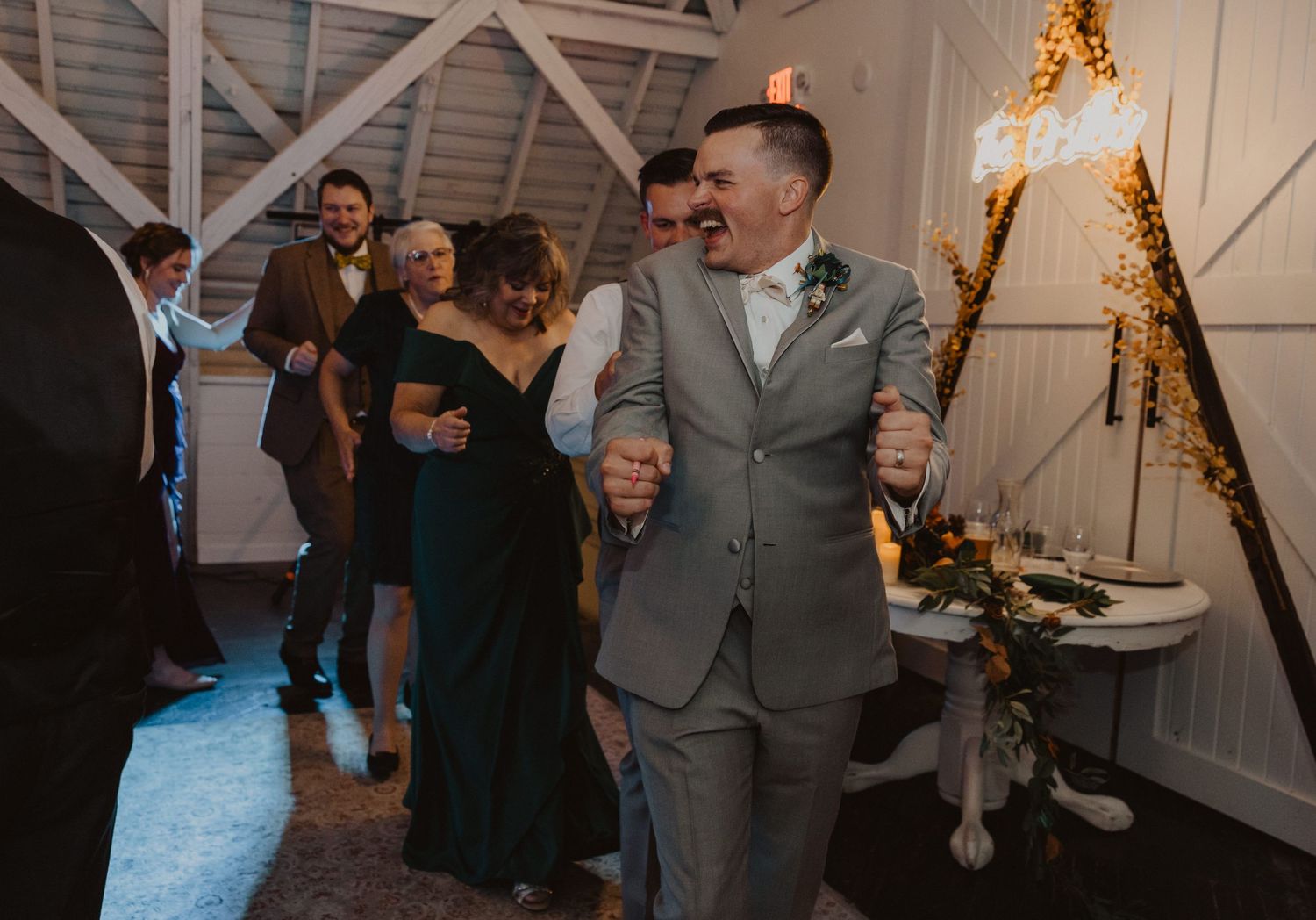 Wedding reception guests dancing and socializing in a rustic barn setting with ambient lighting.