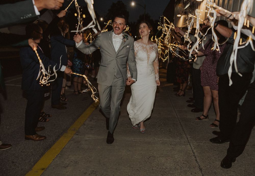 A couple exits their wedding celebration at night while guests wave sparklers creating a magical light tunnel.